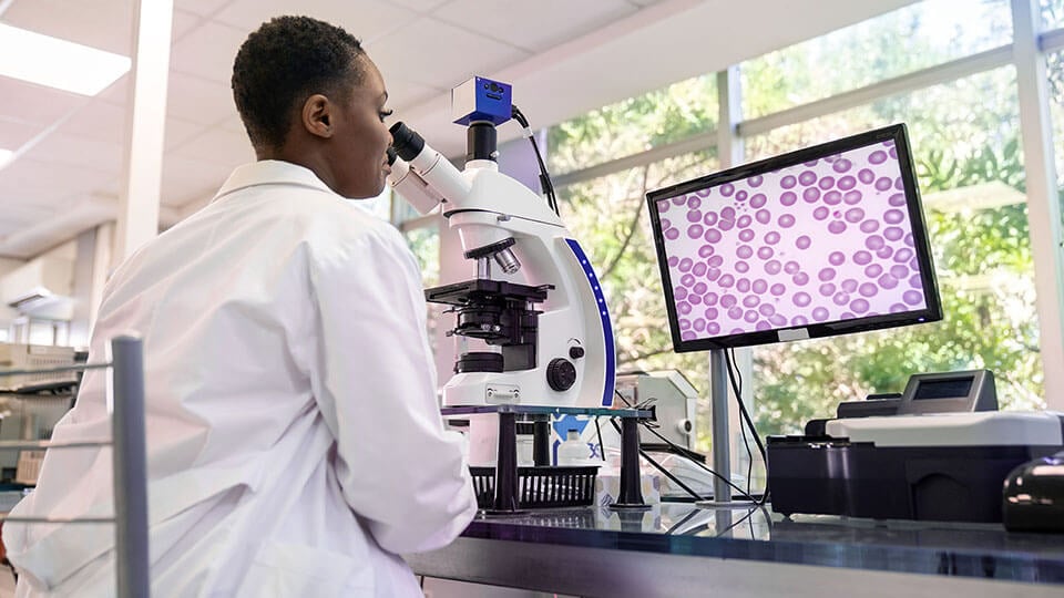 Rear view of a woman pathologist looking at a sample using microscope with magnified image seen on computer screen